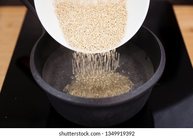 Bowl Pouring Raw Quinoa Grains Into Boiling Water Inside A Hot Black Cooking Pot Heating On A Ceramic Hob On A Kitchen Background. Quinoa Easy  And Healthy Cooking At Home.