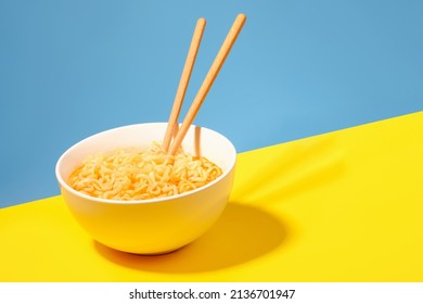 A bowl with ready instant noodles and chopsticks on blue and yellow background. - Shutterstock ID 2136701947