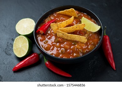 Bowl of salsa sauce with nachos chips, red hot chili peppers and lime, studio shot over black stone background