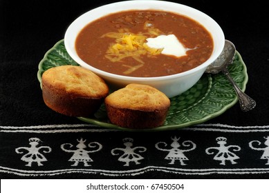 Bowl Of Spicy Tex-Mex Chili And Jalapeno Cornbread With Melted Cheese And Dollop Of Sour Cream On Black Place Mat With Aztec Design.