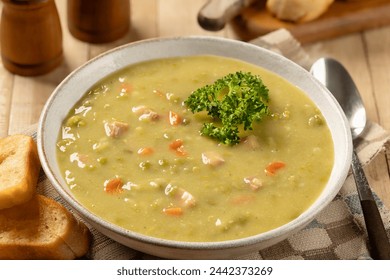 Bowl of split pea soup with ham and carrots garnished with parsley and toasted baguette on a wooden table