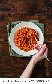 A Bowl Of Stewed Cheak-pea With Hot Tomato Sauce On A Napkin, On Wooden Background, Overhead Shot. A Hand Holding Full Spoon.