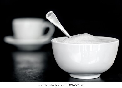 Bowl of sugar with spoon.  Coffee cup behind, over black.  Selective focus.