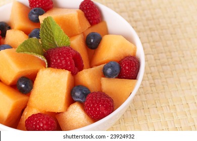 Bowl of summer fruit salad with raspberries, melon, blueberries and mint leaves in white bowl