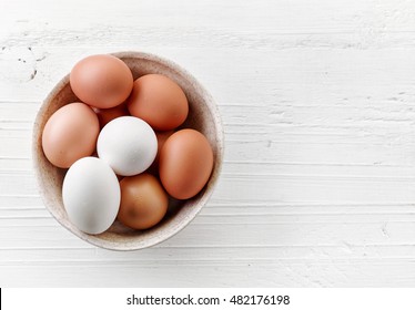 bowl of various fresh eggs on white wooden table, top view