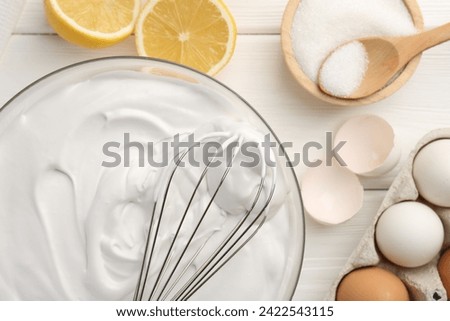 Bowl with whipped cream, whisk and ingredients on white wooden table, flat lay