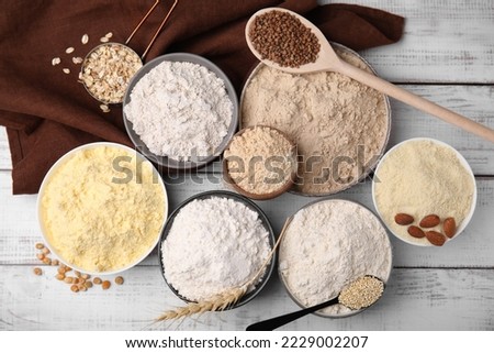 Bowls with different types of flour and ingredients on white wooden table, flat lay