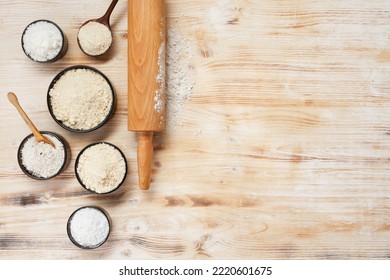 Bowls with different types of flour, wooden spoons and rolling pin on a white and brown wooden table, top view. Space for text.