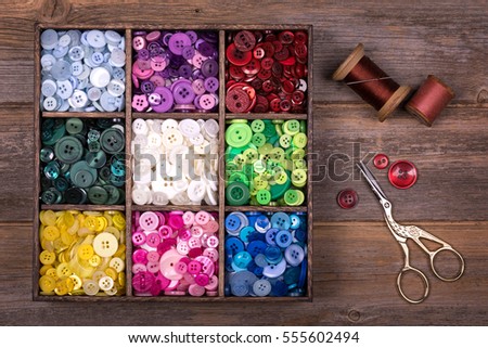 A box of colourful buttons, sorted into colour groups, in an old wood box. Spools of thread, a needle and embroidery scissors are placed to one side. Needlecraft and scrapbooking theme. 