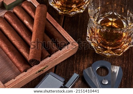 Box with cuban cigars, lighter and cutter on old wooden table top. Two glasses of whiskey or alcohol on the background.