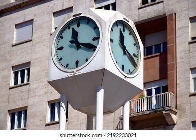 Box Or Cube Shaped Large Painted White Metal Street Clock With Large Black Clock Hands On High Circular Posts. Residential Building With Stone Facade And Balcony In The Background. Urban Setting.