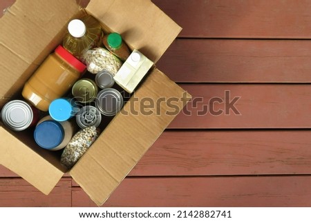 A box of donated canned goods and non-perishable foods for a food pantry for the poor sitting on red, wood panel background.  Copy space.