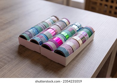 A box full of colorful rolls of washi tape having different patterns, textures and colors on a wooden table. The tapes are ready to be used to decorate an interior, gift or post card, gift.