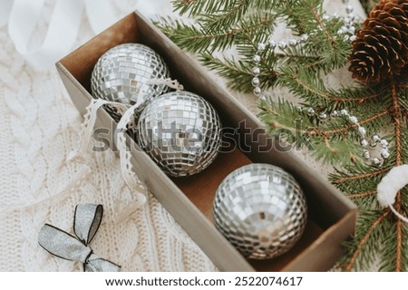 Box with three mirror balls, silver bow, gerlands, ribbons for Christmas tree decoration and fir branch on white knitted background