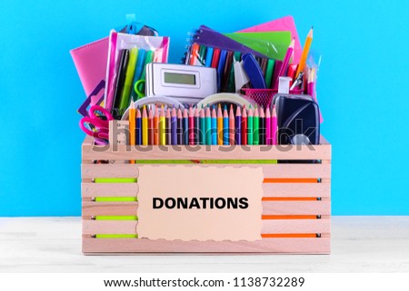 Box with various school and office supplies with a sign on a bright blue background. Donation concept