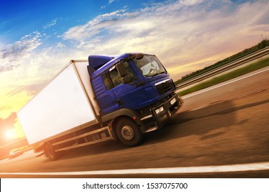 A Boxed Truck Driving Fast On The Countryside Road With Green Trees And Bushes Against A Night Sky With A Sunset