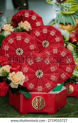 The boxes are presented in a larger, matching red hexagonal wooden tray that also bears a prominent golden Double Happiness emblem on its front. To the left, a lush bouquet of cream and red roses adds a soft, romantic contrast to the geometric lines of the boxes. In the background, a glimpse of a traditional dragon and phoenix sculpture made from fruits and leaves can be seen, which is a hallmark of high-end Vietnamese ceremonial decor. The dominant use of red and gold symbolizes luck, wealth, and prosperity for the couple.