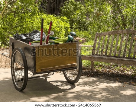 Boxy handcart with watering cans, a bucket, and other equipment, parked by a vacant wooden bench, in dappled sunlight along a paved path in a botanical garden on a spring morning, southwest Florida