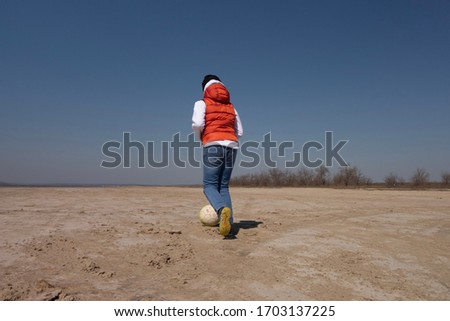 A boy of 10 years old in a white sweatshirt and orange vest plays football on a deserted beach in solitude.