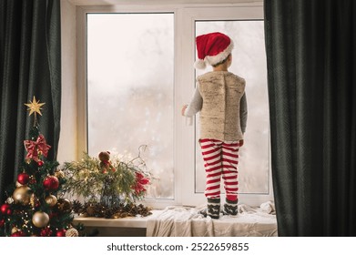 boy 3-4 years old in red and white stripped pajamas and Santa Claus hat plays on the window near the decorated New Year tree with lanterns and gingerbread house. Christmas symbols, holiday card - Powered by Shutterstock