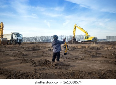 a boy of 3-4 years old in warm clothes at a construction site with his back in the frame holds a toy excavator and looks at the large construction equipment in front of him. Little builder