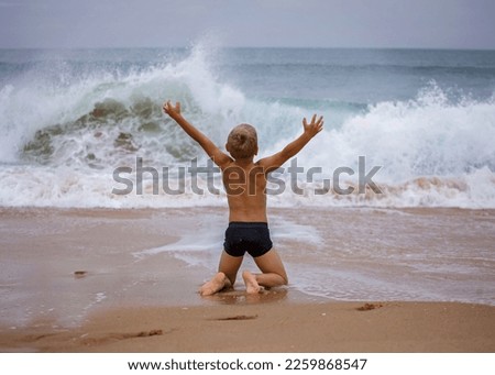 A boy of 7-8 years old sits on the ocean shore with his arms open towards the wind and waves. Storm on the ocean in summer. The concept of freedom, courage and challenge. Place for text