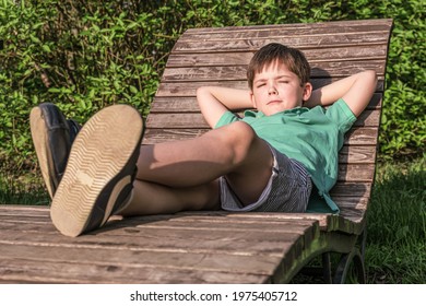 Boy Of 8 Years In T-shirt And Shorts Lies On Wooden Deck Chair In The Sun Against Background Of Green Leaves. The Boy In Relaxed, Lazy Position, Narrowed His Eyes.