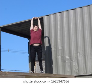 Boy About 20 Years Old, Blond, With Short Hair, Standing Next To Containers, With Arms Up Doing Stretching.