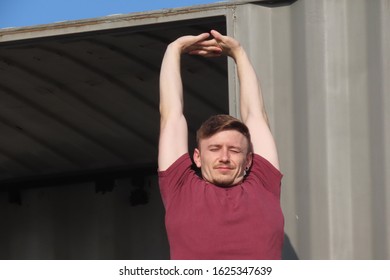 Boy About 20 Years Old, Blond, With Short Hair, Standing Next To Containers, With Arms Up Doing Stretching.