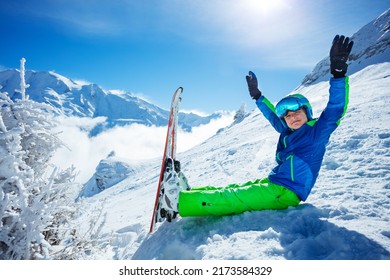Boy with alpine ski sit in snow turn to camera smile lifting hands, wear helmet and sport glasses on top of the mountain over peaks covered by clouds on background