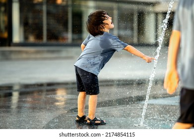 a boy of Asian appearance with long hair in a blue T-shirt is bathing in a fountain on a hot summer day. The kids are having fun . The boy cools down from the heat with the water of the city fountain - Shutterstock ID 2174854115