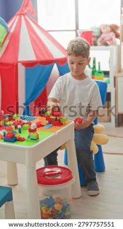 Boy assembling colorful blocks on a play table by a red striped play tent in a building; creative concentration.