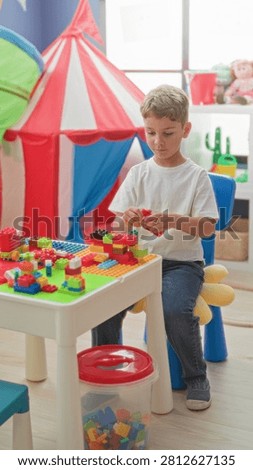 Boy assembling colorful blocks on a play table by a red striped play tent in a building; creative concentration.