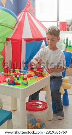 Boy assembling colorful blocks on a play table by a red striped play tent in a building; creative concentration.