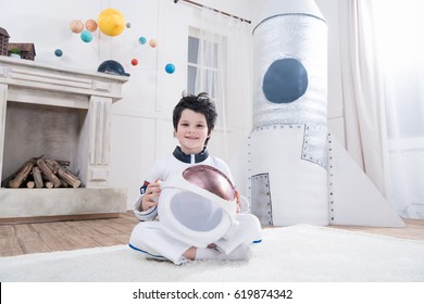 boy in astronaut costume holding helmet, toy rocket behind 