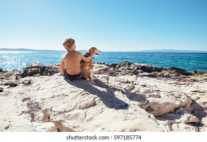 Boy with beagle dog sitting together on rocky sea coast at sunny afternoon