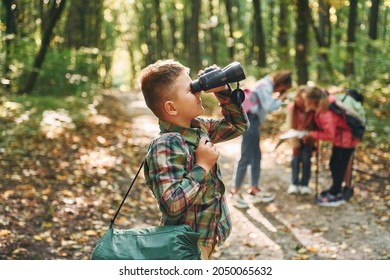 Boy With Binoculars Standing In Front Of His Friends. Kids In Green Forest At Summer Daytime Together.