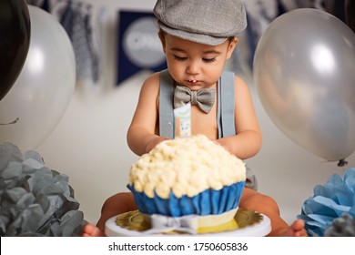 Boy Birthday Cake Smash. cute one year old boy with dark hair, in shorts with suspenders sits on a gray background near the balloons