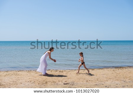 A boy in a black speedo running to mom brunette in a white dress on the coast