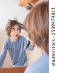 A boy is blow drying his hair in front of a mirror. He is wearing a blue shirt and has a pocket on the front