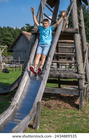 A boy in a blue t-shirt and denim shorts slides down a wooden playground slide, arms raised with exhilaration. Perfect for themes of joy, outdoor play, and childhood adventures. High quality photo