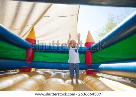 Boy bouncing inside brightly colored inflatable bounce castle under beige shade canopy in backyard. Playground, recreation, vibrant, outdoor, childhood, activity, safety