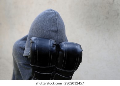 Boy with boxing gloves covering his face. Young boxer training. Boy in a hooded sweatshirt and boxing gloves practicing. Contact sport. Training. Light background with empty space. Fight. Violence. - Powered by Shutterstock