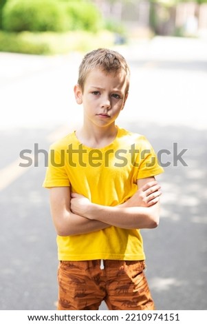 A boy in a bright yellow T-shirt stands with his arms crossed over his chest and stares earnestly into the camera. vertical photo