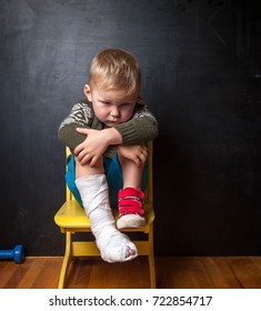 Boy With Broken Leg On Black Background. Child In Plaster Cast Sitting On The Chair. Sad And Unhappy Child On Small Chair. Upset Toddler Boy. 