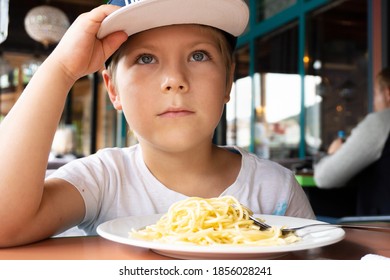 A Boy In A Cap 6 Years Old Is Sitting In A Cafe And Does Not Want To Eat Spaghetti, Looking Directly At The Camera