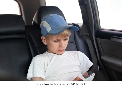 A Boy In A Cap Sits In The Back Seat Of The Car, Looks At The Phone Screen With Enthusiasm And Concentration.