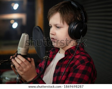 Boy in check shirt and headphone is singing or talking into microphone with pop filter in voice recording studio. Young singer recording his voice for phonogram performance for entertainment. 