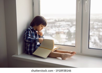 Boy Child Sits On The Window Of A House With A Book In Winter On A High Floor