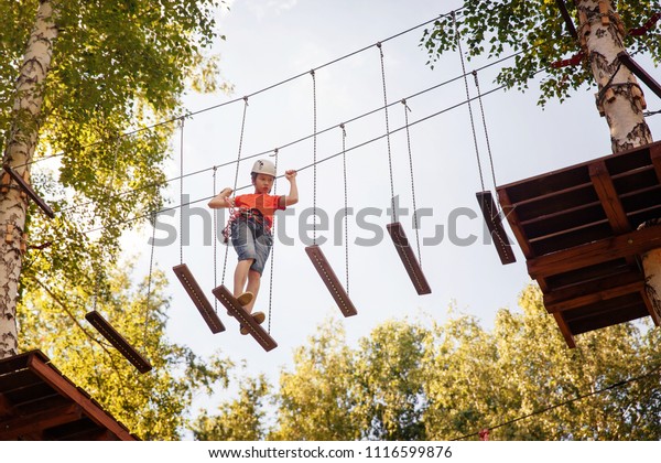 Boy with climbing gear in an\
adventure park are engaged in rock climbing or pass obstacles on\
the rope road. Rope park in pine wood forest against the\
sky
